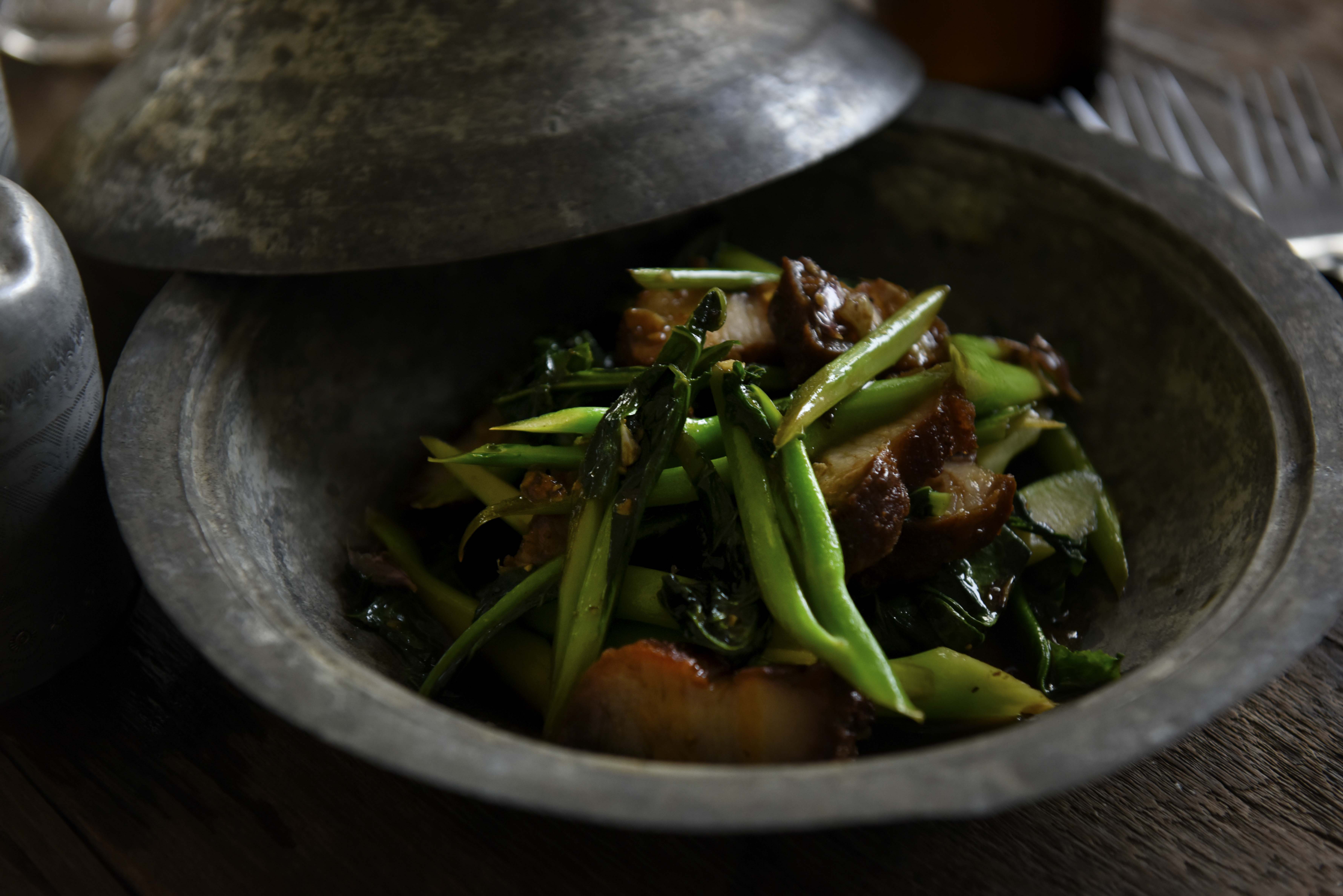 Editorial food photograph — braised pork and greens in a stone bowl, dramatic low light
