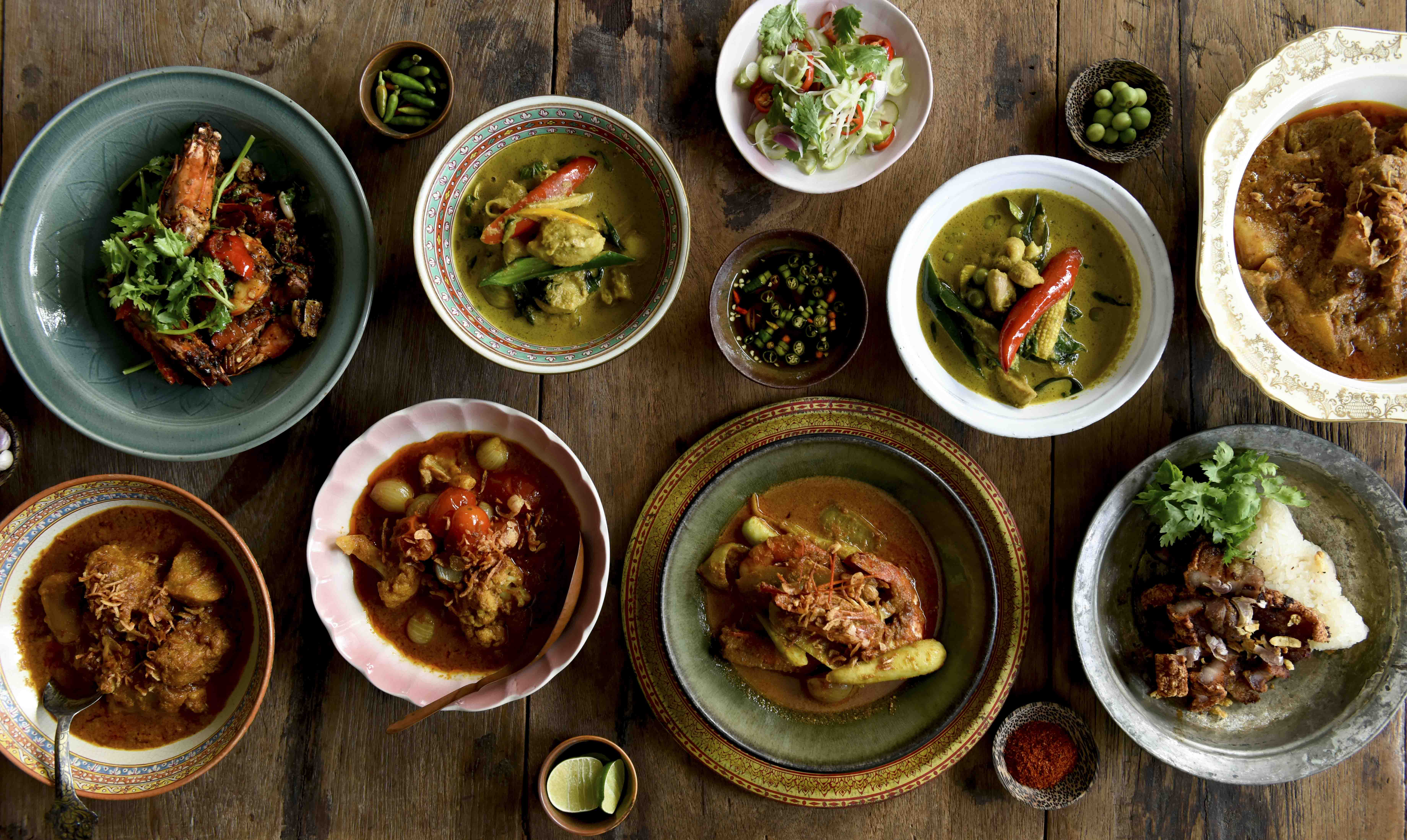 Editorial overhead — a Thai meal spread across a worn wooden table, curries, salad, and condiments in mismatched bowls
