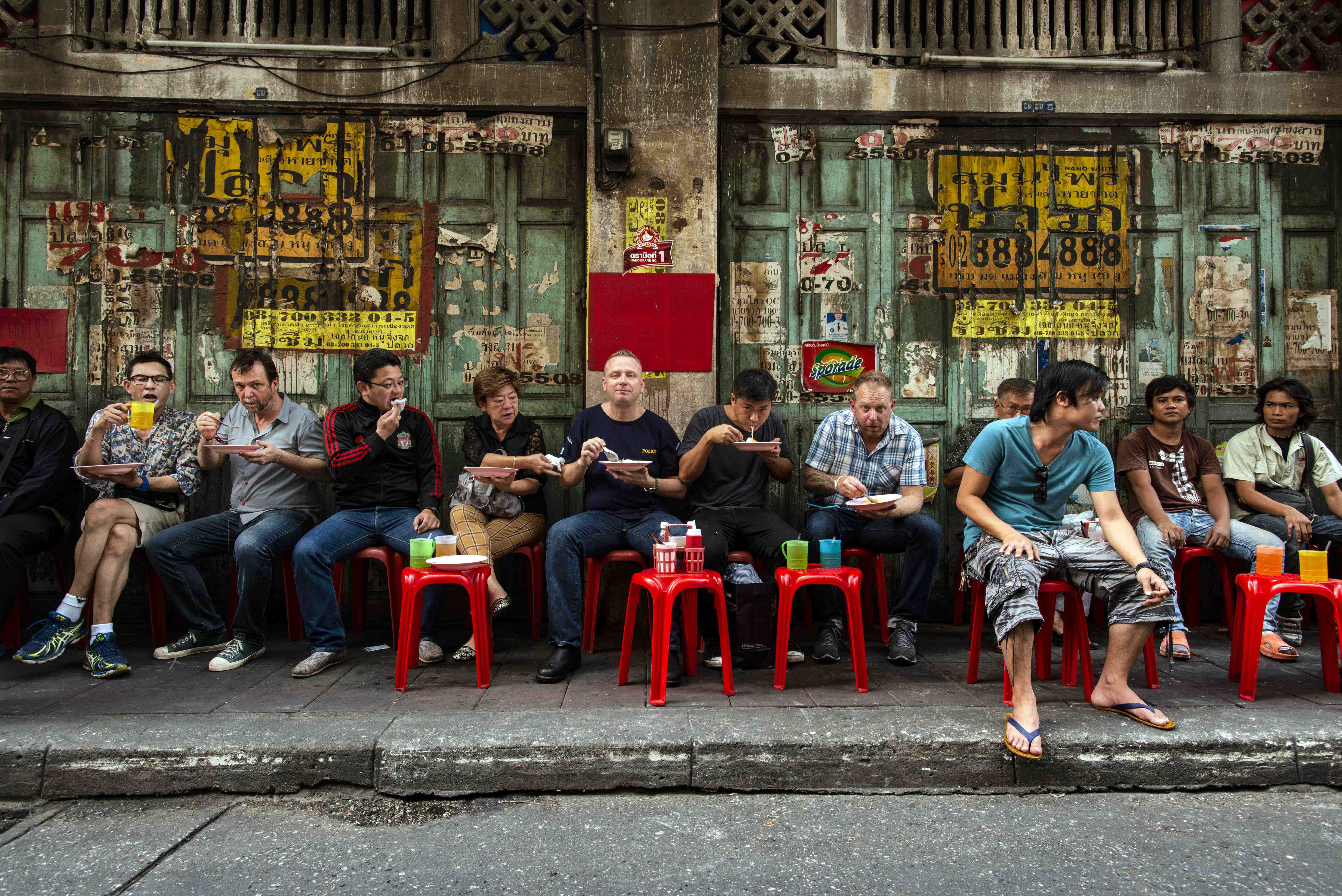 Bangkok street food — chefs and locals sharing a row of red plastic stools on a Yaowarat sidewalk, weathered shophouse door behind